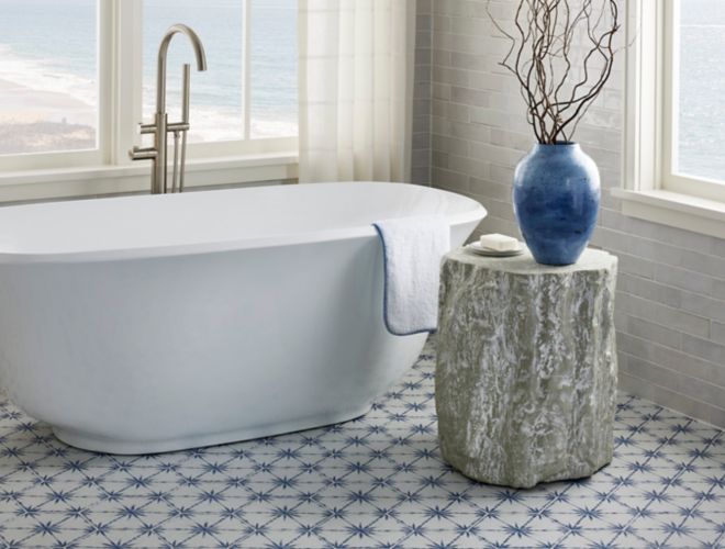 White tub in a bathroom with large windows and blue and white porcelain hexagon tile on the floor. Small table with decorative branches in a blue vase and soap.