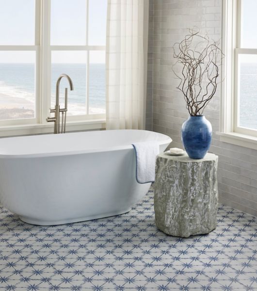 White tub in a bathroom with large windows and blue and white porcelain hexagon tile on the floor. Small table with decorative branches in a blue vase and soap.