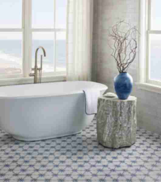 White tub in a bathroom with large windows and blue and white porcelain hexagon tile on the floor. Small table with decorative branches in a blue vase and soap.