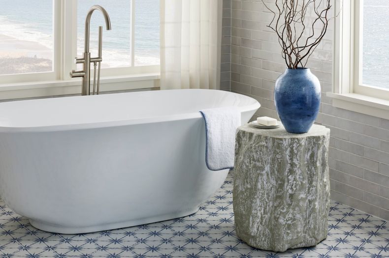 White tub in a bathroom with large windows and blue and white porcelain hexagon tile on the floor. Small table with decorative branches in a blue vase and soap.