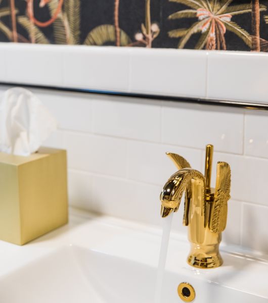 Bathroom sink with gold fixture and tissue box. White tile and black wallpaper with palm trees and monkey.