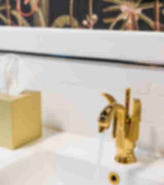 Bathroom sink with gold fixture and tissue box. White tile and black wallpaper with palm trees and monkey.