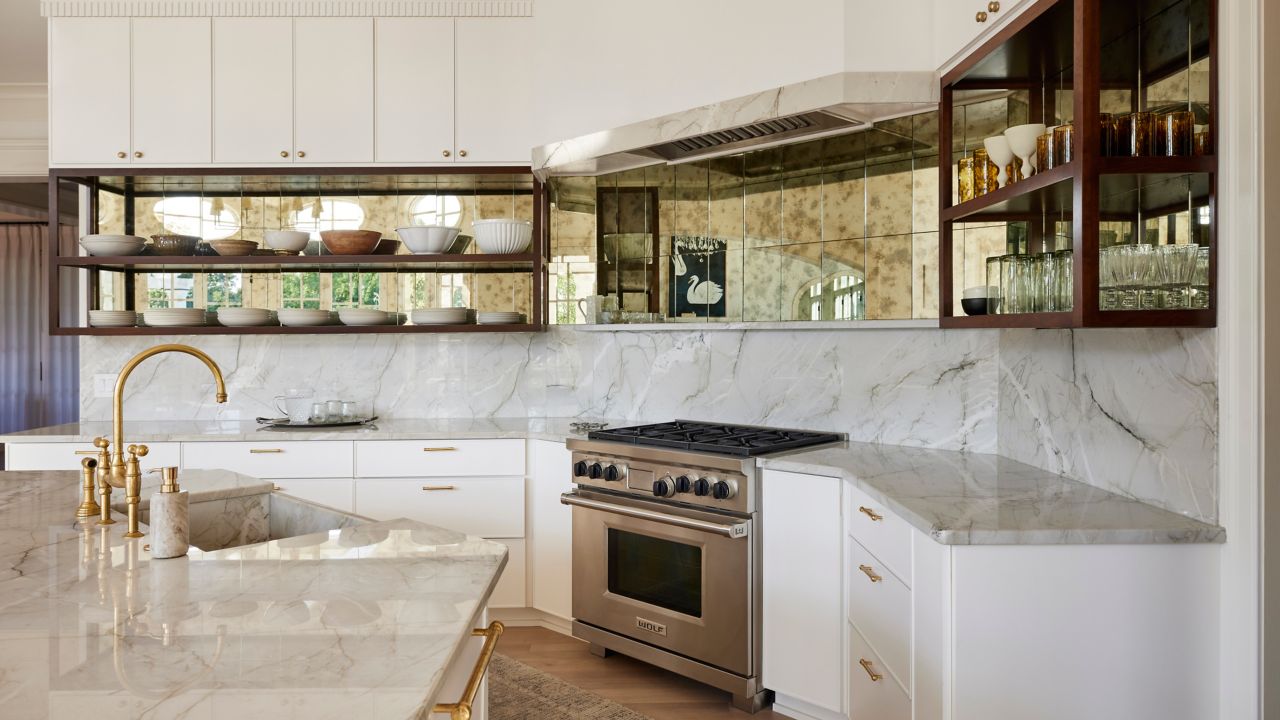 A bright, modern kitchen with marble countertops, wood flooring, a large format marble tile backsplash, and reflective gold patina tiles above the backsplash.