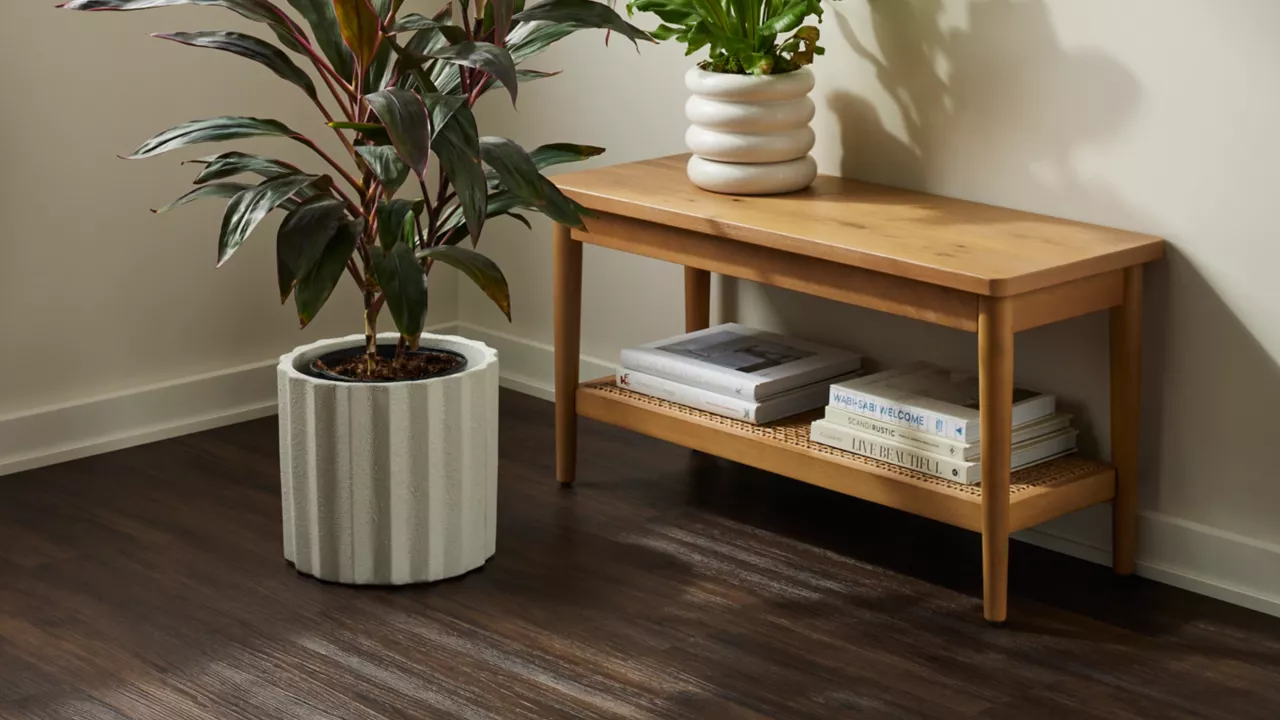 
         A wooden bench with books on a shelf sits on a dark brown luxury vinyl plank floor, next to two potted plants in white ribbed planters.