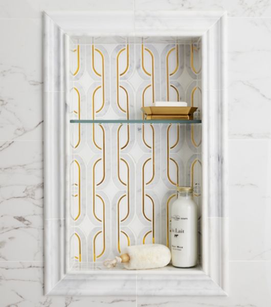 Close up view of shower niche with grey and white profiles and marble mosic with gold color accent design. Shower wall is also a large marble look in grey and white.  Glass shelf and bathing accessories in niche.