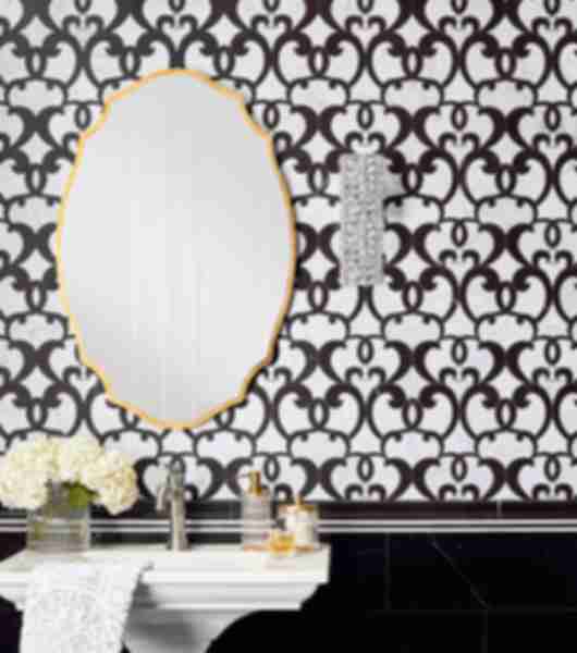 White bathroom sink with soaps and flowers. Gold-frame oval mirror on a black and white tile wall.
