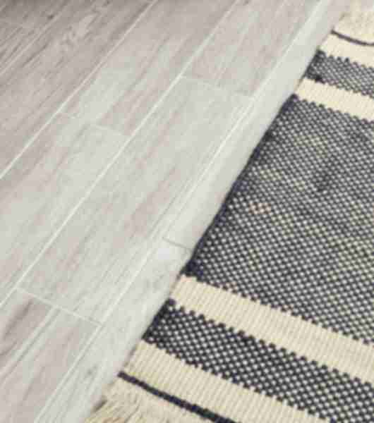 Floor shot of 12x24 inch rectangular wood look porcelain tile in matte finish. A woven rug accents the floor.
