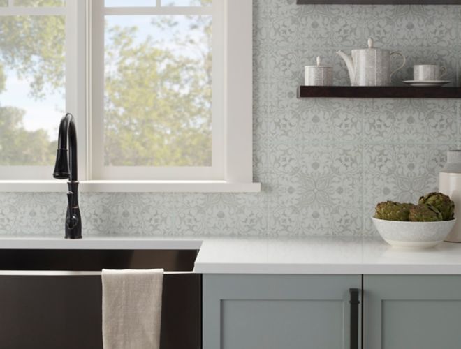 This farmhouse-style kitchen features gray lower cabinets, white countertops, and a dark under-mount sink and faucet. The wall above the sink is covered in square porcelain tile with a delicate floral pattern in shades of cream and weathered green. Open-style wall shelving adds an additional farmhouse touch.