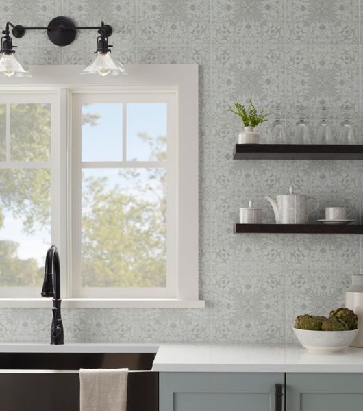 This farmhouse-style kitchen features gray lower cabinets, white countertops, and a dark under-mount sink and faucet. The wall above the sink is covered in square porcelain tile with a delicate floral pattern in shades of cream and weathered green. Open-style wall shelving adds an additional farmhouse touch.