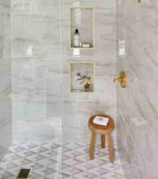 Walk-in shower with walls covered in large-format, marble-look porcelain tiles that combine shades of cream and tan. Gold trim pieces frame out the recessed wall niches, and match the gold-tone shower hardware.