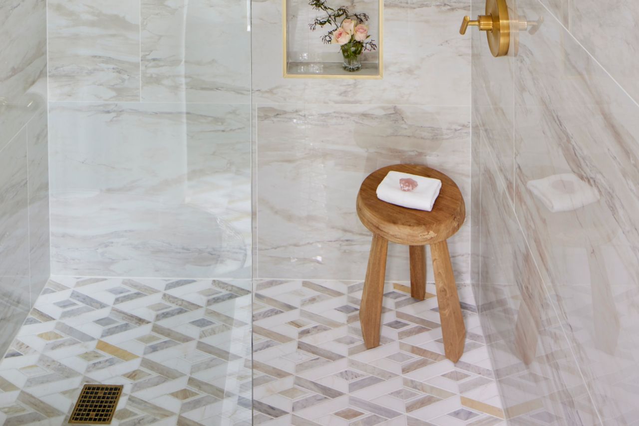 Walk-in shower with walls covered in large-format, marble-look porcelain tiles that combine shades of cream and tan. Gold trim pieces frame out the recessed wall niches, and match the gold-tone shower hardware.