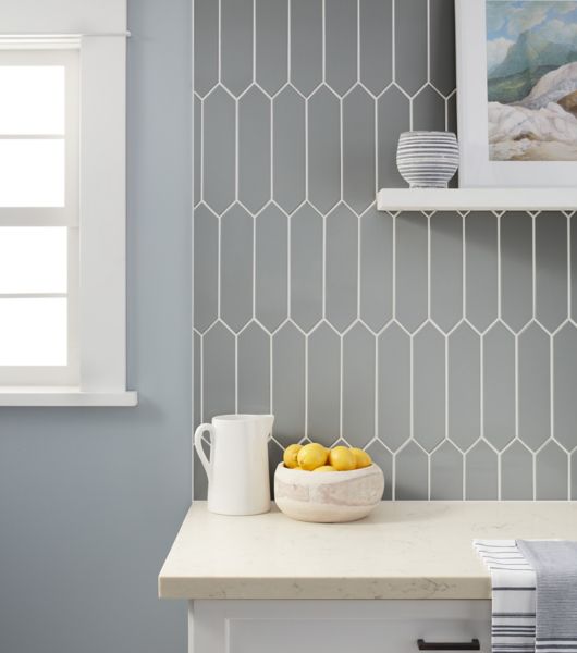 Kitchen backsplash using cool greige toned tile in a picket shape ran vertically.  Countertops are light beige and cabinets are white with black hardware.