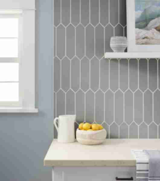 Kitchen backsplash using cool greige toned tile in a picket shape ran vertically.  Countertops are light beige and cabinets are white with black hardware.