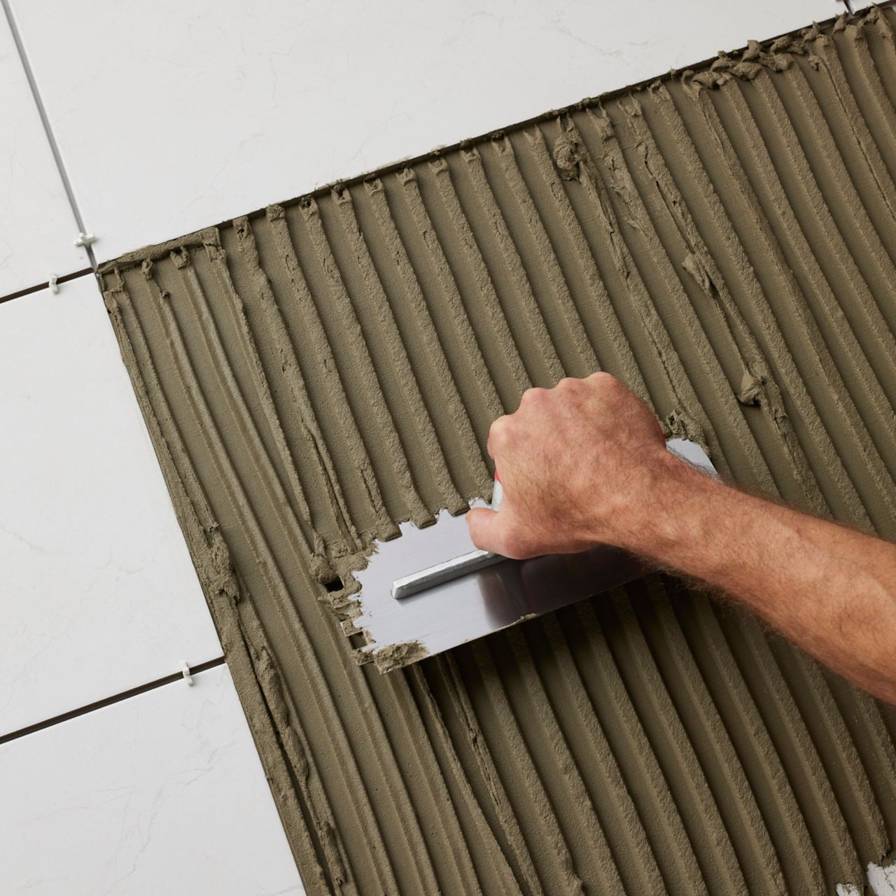 A hand holding a square notched trowel spreading thinset surrounded by white tiles.