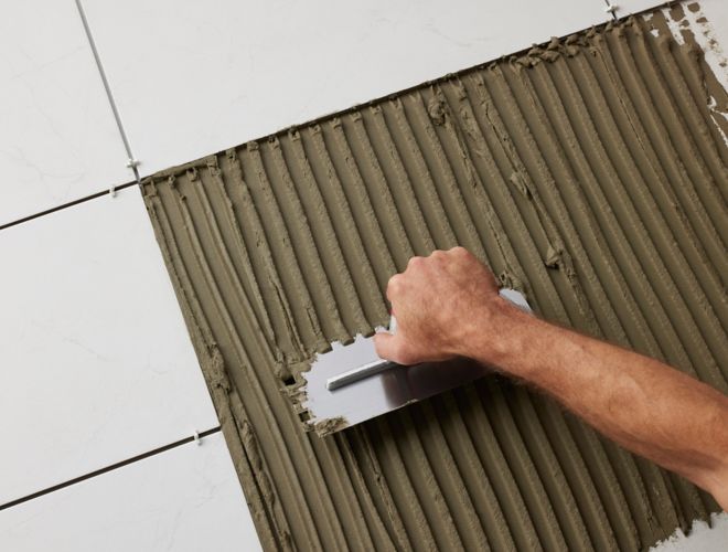 A hand holding a square notched trowel spreading thinset surrounded by white tiles.