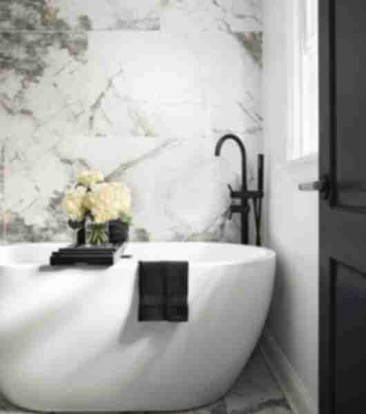 Large-format marble-look porcelain tile in shades of white and gray covers the walls and floor of a bathroom that also features a deep white soaker tub with matte black faucet.