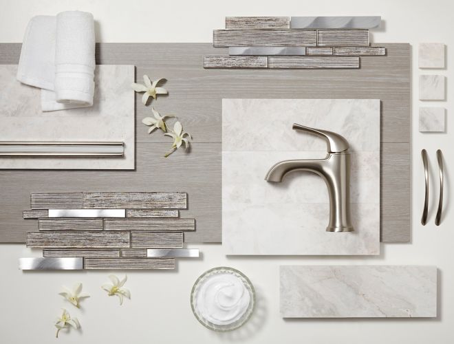 Glass and metal mosaics, grey faux wood tile, white marble tile, and trim pieces lay on a table top with a soft towel, face cream, and flowers.