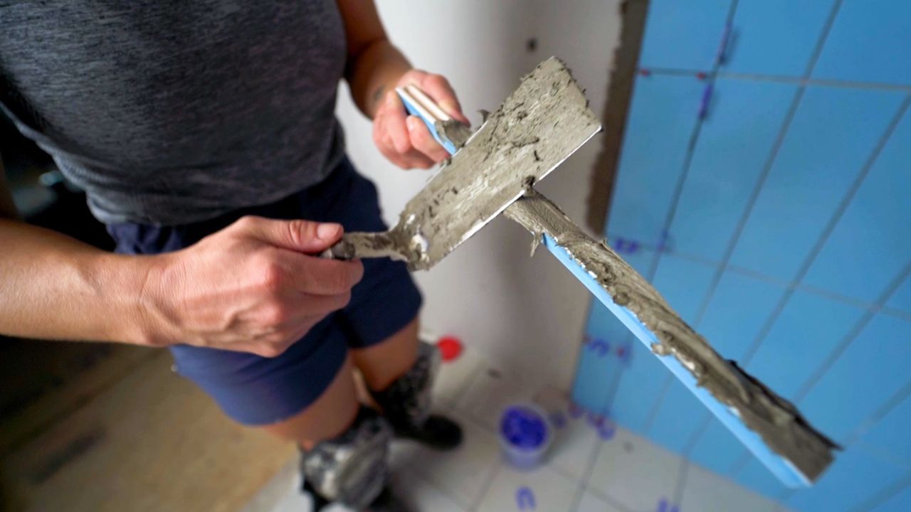 A person using a trowel to apply thinset mortar to the back of a piece of pencil trim tile. The channel on the back of the tile has been completely filled with mortar so the piece will securely bond to the wall.
