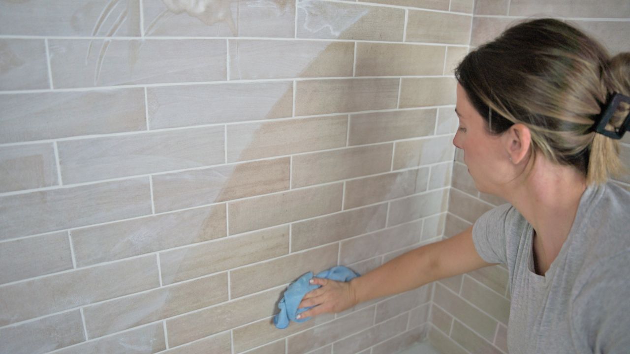 A tile pro wipes the grout haze off of a newly installed tile shower. You can see a clear different between the tile she has wiped clean, and the haze on the rest of the tile.