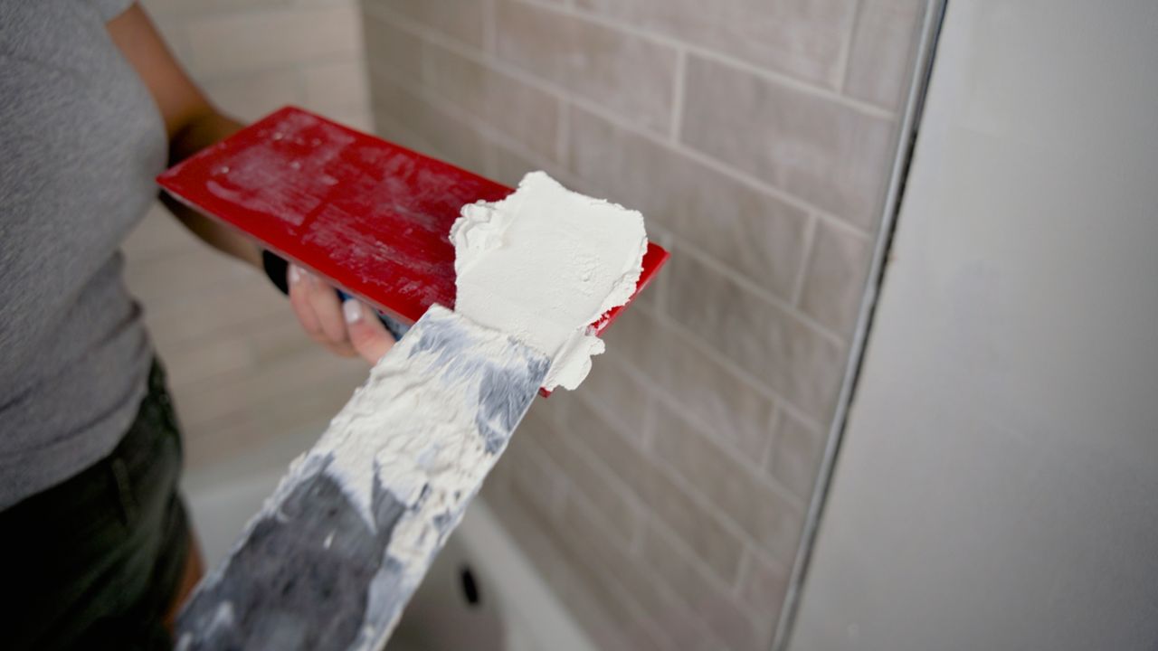 Using a margin trowel to add grout to a grout float, in preparation of grouting a newly tiled shower wall.