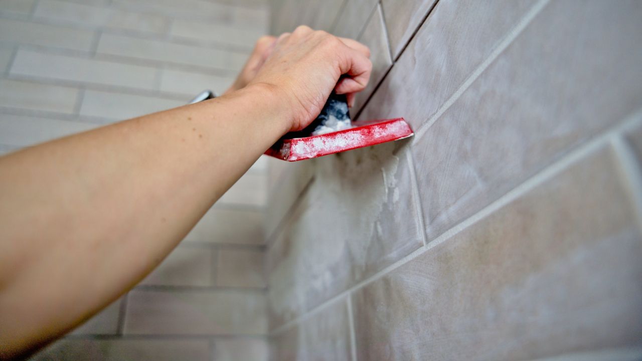 Using the long side of a grout float to fill grout joints and wipe away the majority of excess grout from a tiled shower wall.