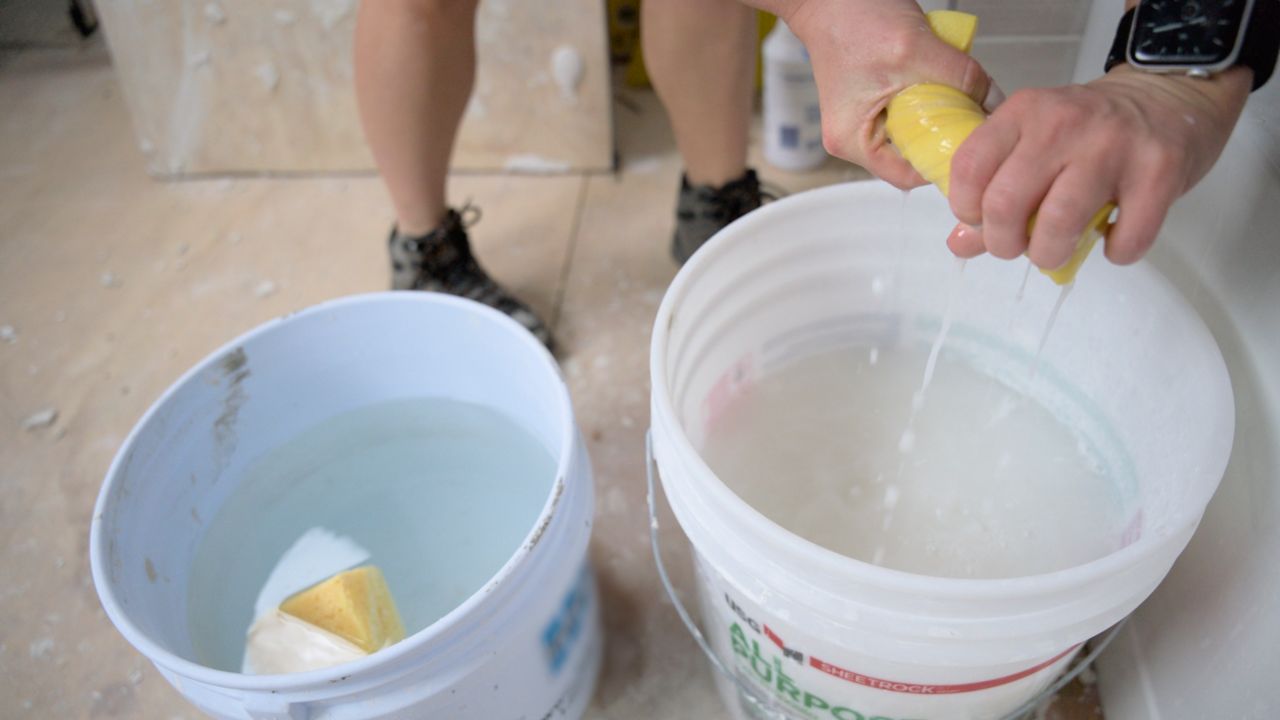 A tile pro demonstrates her method for using two buckets of water to quickly and efficiently wipe remaining grout haze from the surface of a newly tiled shower wall.
