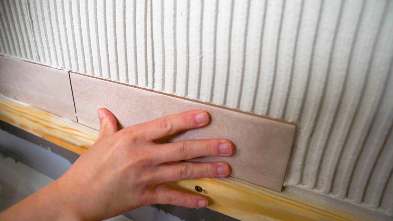 A tile pro applies uses her hands to apply pressure to a tile she is installing. Pressing the tile and moving it back and forth slightly helps to collapse notches in the thinset behind it, removing air and promoting a strong bond between the tile and the wall.