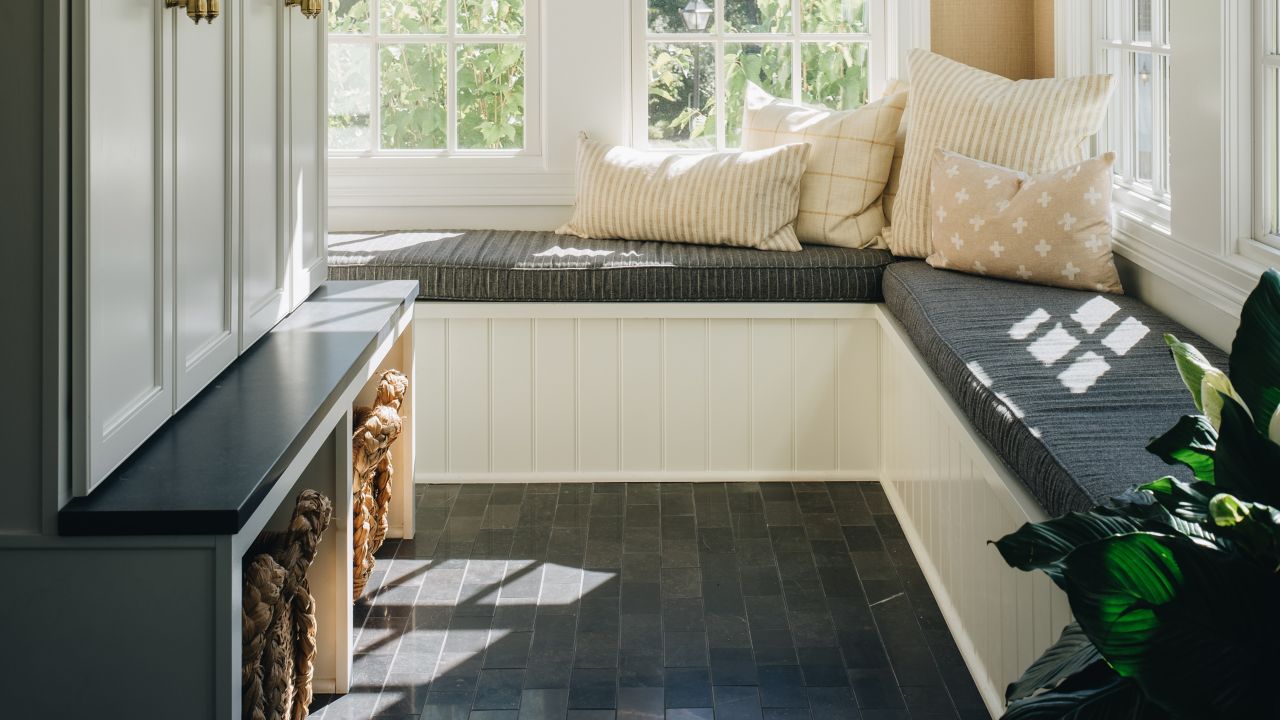 A mudroom with beige walls and a white slatted ceiling has a built-in white bench with a dark gray cushion and several throw pillows. The floor is covered in dark, rectangular tiles, and a white cabinet with gold handles stands to the left.