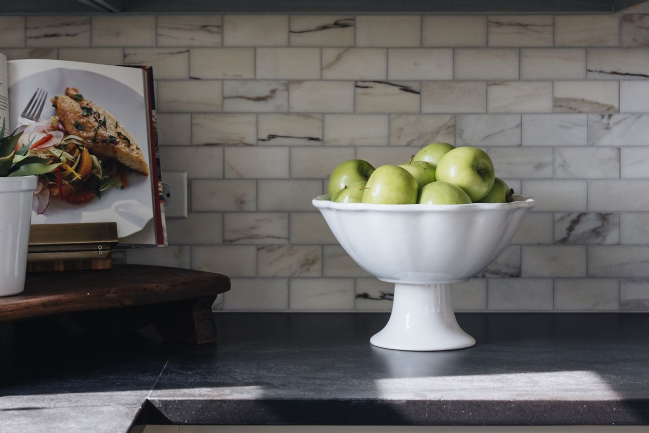 A kitchen with a marble subway tile backsplash and a bowl of fruit on the black counter.