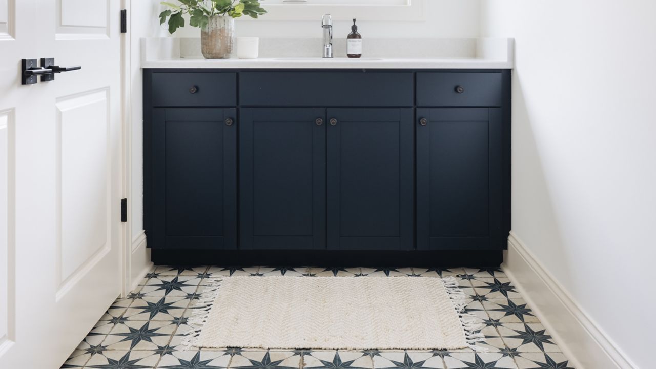 
A navy blue cabinet with a white countertop and small white sink sits in a white room with a window and a white door with black hardware, on a patterned tile floor with a blue and white starburst design.