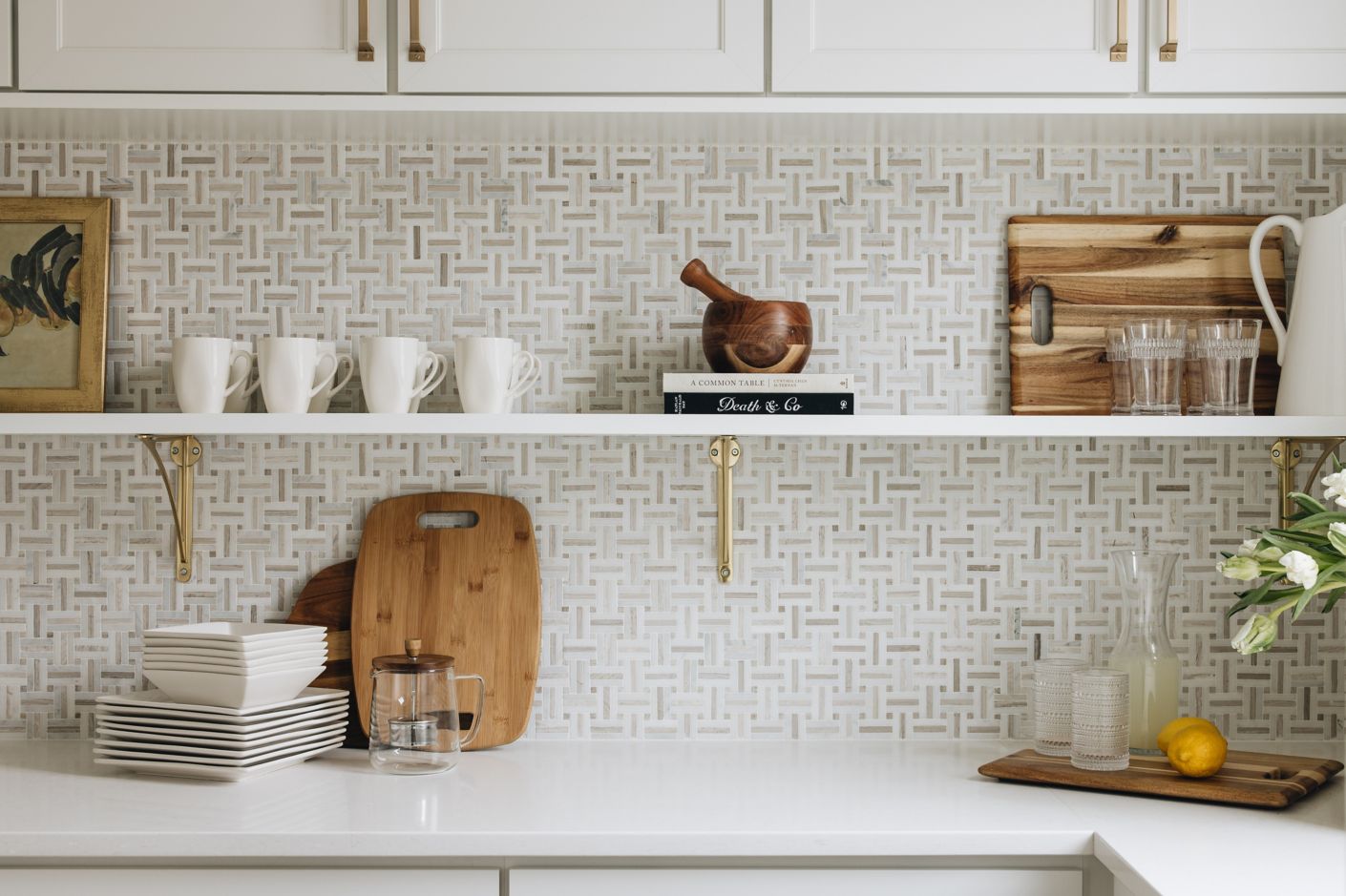 Kitchen counter and shelf with cups, cutting board and other cooking elements before white and beige tile.