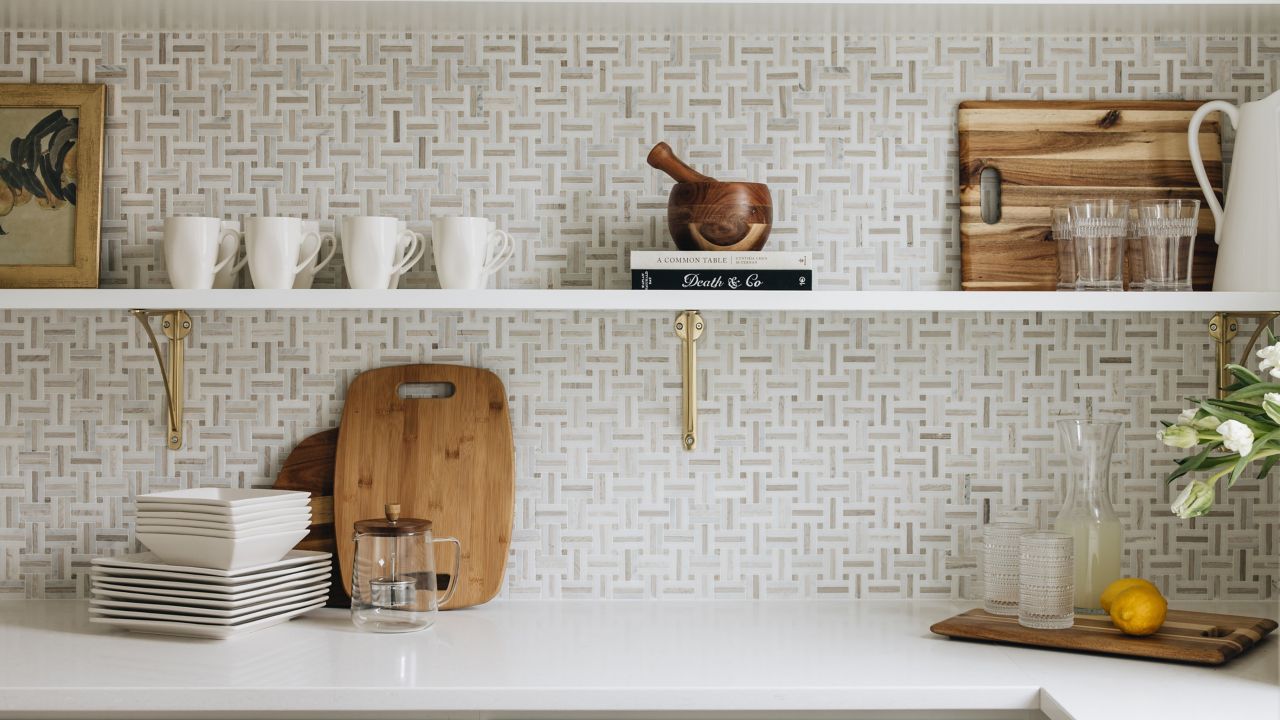 A view of a kitchen countertop, backsplash, and cabinets. The kitchen has white kitchen countertops, white shelves mounted on the backsplash, and white cabinets above. The tile backsplash is a mosaic pattern of white and brown tiles