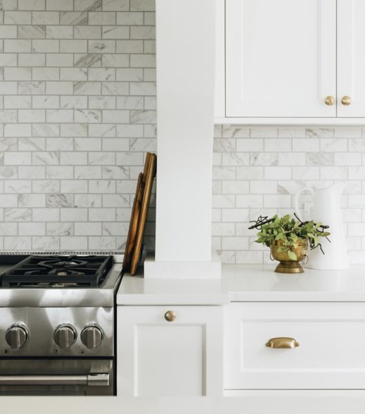 The white cabinets, gold hardware and white counters in this kitchen complement the marble subway tile backsplash above the counters and stove. The marble is white with soft gray veining.