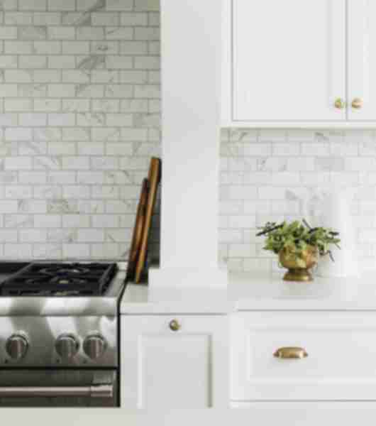 The white cabinets, gold hardware and white counters in this kitchen complement the marble subway tile backsplash above the counters and stove. The marble is white with soft gray veining.