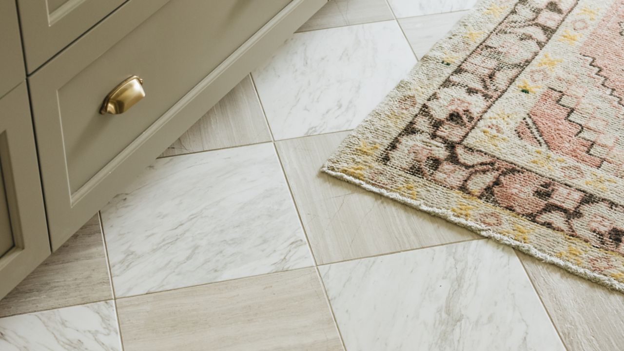 
A beige runner rug with pink and yellow accents sits on a checkerboard floor of white marble and beige wood-grain tiles, in front of a light gray cabinet with gold hardware.