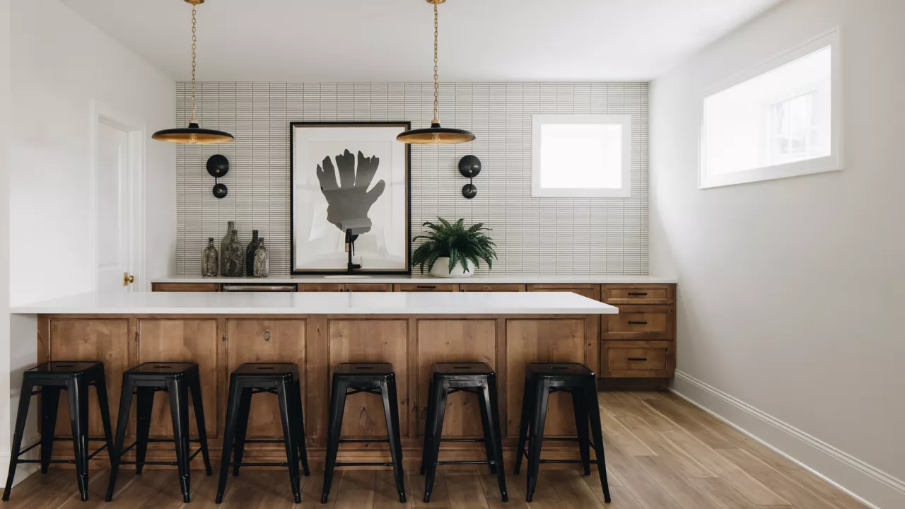 
A long wooden kitchen island with a white countertop and six black bar stools sits in front of a wood-paneled wall with white tile and a large black and white art piece.
