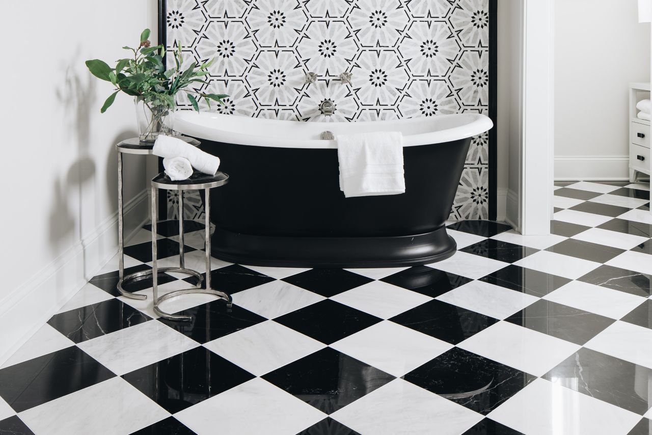 Bathroom with black and white checkerboard floor tile and a black and white mosaic accent wall. Bathtub and small table with a plant.