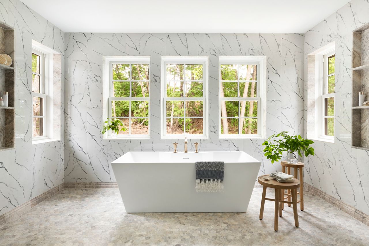 This bright, spacious bathroom features white marble-look wall tile with grey veining and hexagon-shaped travertine floor tile. 