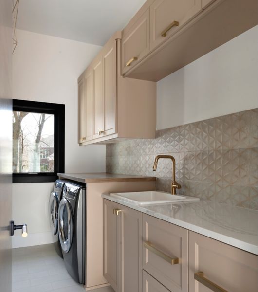 The wall over the sink in a laundry room features a cement-look tile backsplash made up of large-format tiles in neutral tan and grey tones.