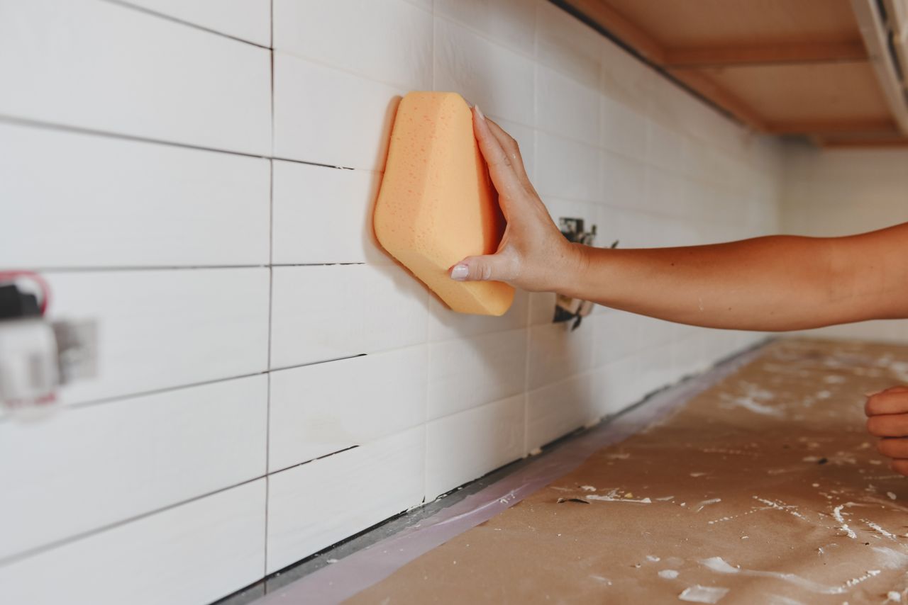 Using a damp sponge to clean excess grout from the surface of a subway tile backsplash before allowing grout to dry.