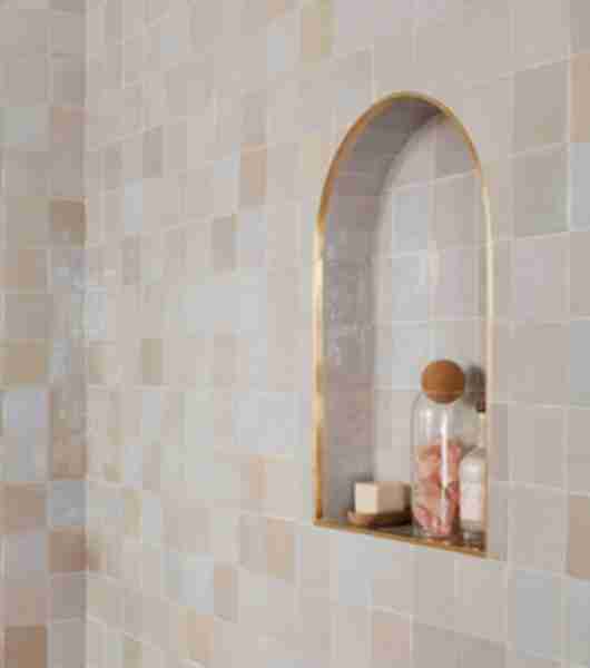A bathroom shower wall and recessed storage niche feature Zellige tile. The niche is arch-shaped and framed in gold-tone trim.