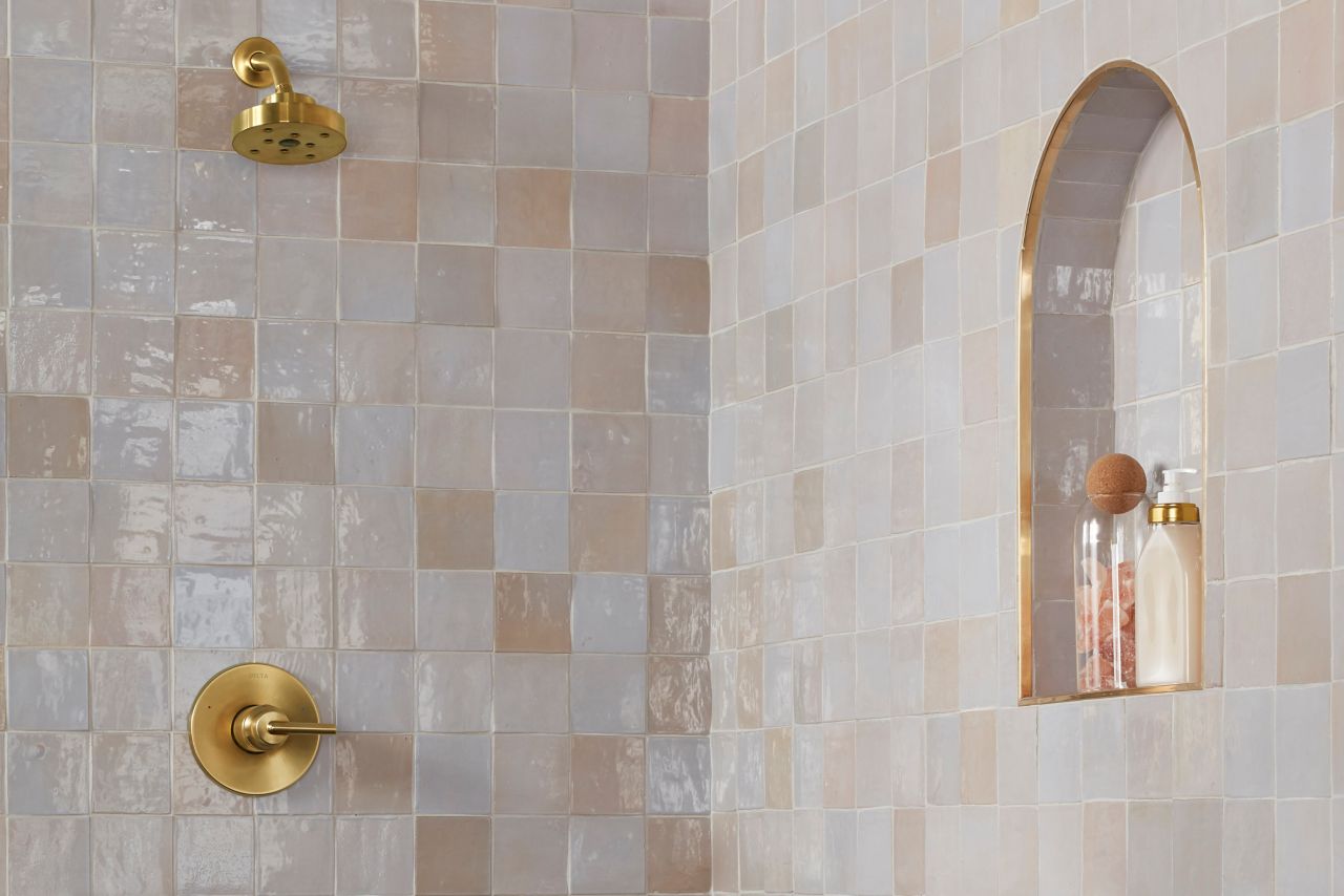 Shower with beige- and pink-toned square handmade tile wall, gold fixtures and small arched shower niche.