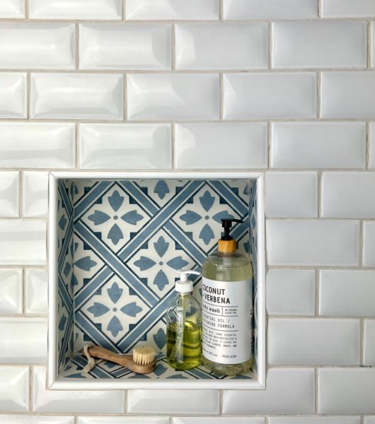 This charming blue and white bathroom features shower walls covered in 3x6-inch white subway tile with a raised, curved surface, bordered in 3x12-inch smokey blue subway tile. The recessed niche is accented with porcelain tile featuring a geometric blue and white floral design.