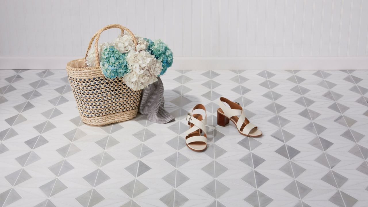 
A woven tote bag filled with light blue and white hydrangeas sits on a light gray and white geometric tile floor, next to a pair of white strappy sandals.