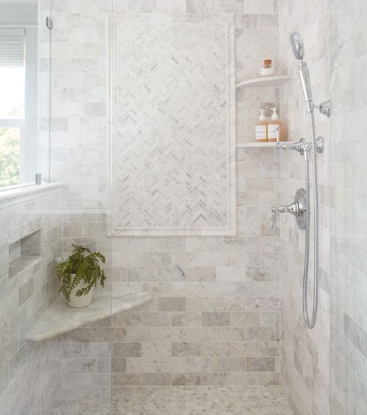 This walk-in shower features light gray marble tiles on the floor and walls, including a built-in corner bench and rounded corner wall shelves to hold personal care items.
