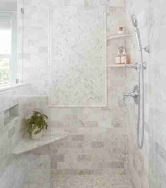 This walk-in shower features light gray marble tiles on the floor and walls, including a built-in corner bench and rounded corner wall shelves to hold personal care items.