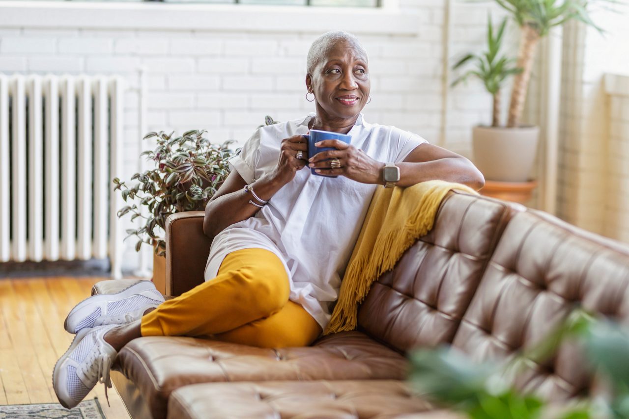 Vibrant senior black woman relaxing at home. She is sitting on the sofa in her living room drinking hot tea or coffee. She is smiling and looking out the window. Retirement, independence, and simple pleasures concepts.