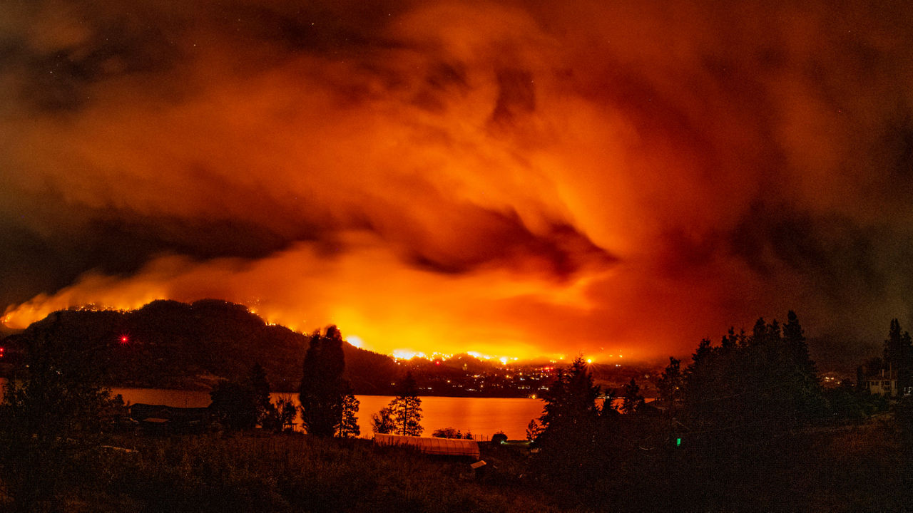 Wildfire burning across hills at night, with an orange smoke‑filled sky glowing above a lake and nearby community.