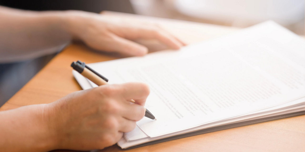 Personne signant un document à l’aide d’un stylo sur un bureau.
