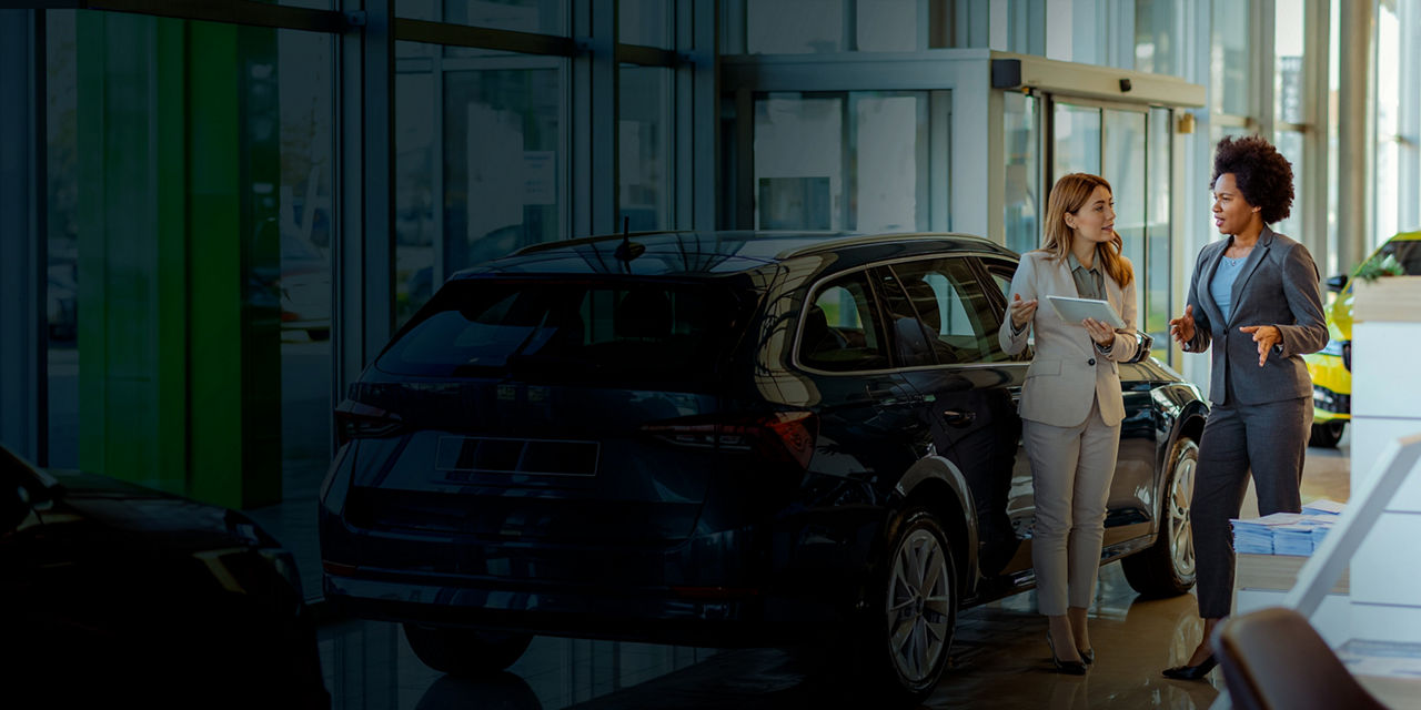 Two women have a conversation while standing beside a blue SUV inside a car dealership.